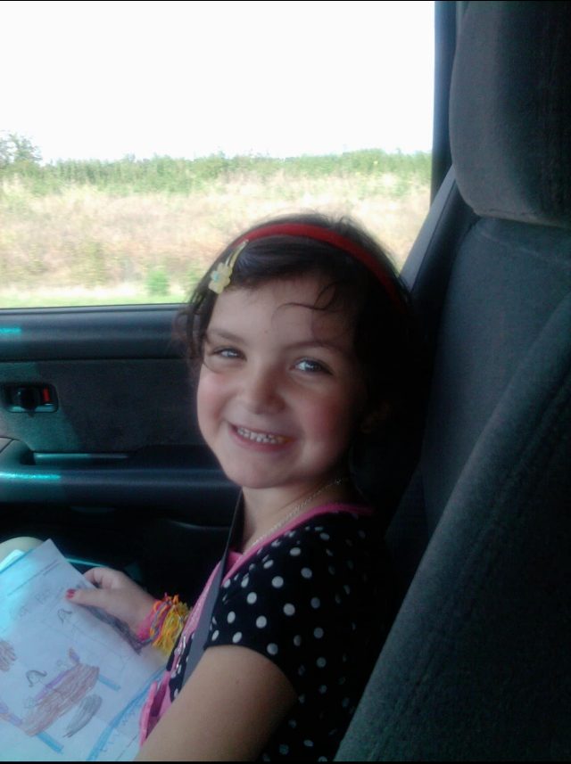 Daryn as a kid, grinning in the back seat with her crayon drawings